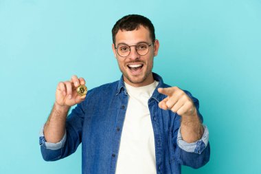 İzole edilmiş mavi arka planda Bitcoin tutan Brezilyalı adam şaşırmış ve ön tarafı işaret ediyor.