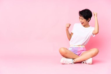 Kısa saçlı genç Vietnamlı kadın, izole edilmiş pembe arka planda oturmuş zaferi kutluyor.