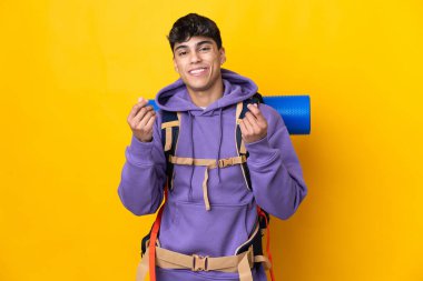 İzole edilmiş sarı arka planda büyük bir sırt çantası olan genç dağcı para hareketi yapıyor.