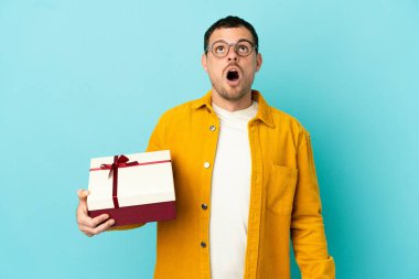 Brezilyalı bir adam mavi arka plan üzerinde bir hediye tutuyor yukarı bakıyor ve şaşırmış bir ifadeyle
