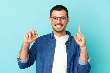 Brezilya 'lı bir adam mavi arka plan üzerinde Bitcoin tutuyor ve en iyinin işareti olarak parmağını kaldırıyor.