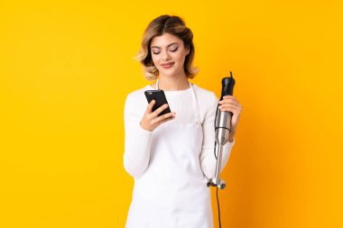 El blenderi kullanan kız sarı arkaplanda izole edilmiş ve cep telefonuyla bir mesaj gönderiyor