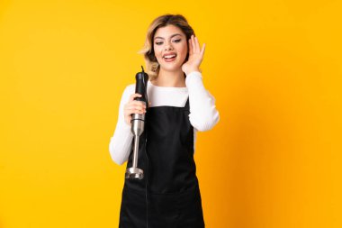 El blenderi kullanan kız sarı arka planda izole edilmiş bir şekilde elini kulağına koyarak bir şeyler dinliyor.