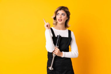 Sarı arka planda izole edilmiş el blenderi kullanan kız mutlu ve yukarı bakıyor