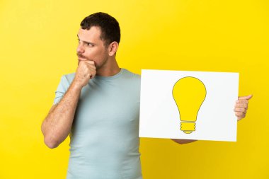 İzole edilmiş mor arka planı olan Brezilyalı bir adam elinde ampul simgesi olan bir levha tutuyor ve düşünüyor.