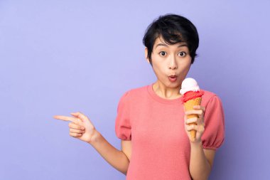 Kısa saçlı genç Vietnamlı kadın izole edilmiş mor arka planda bir kornet dondurma tutuyor. Şaşırmış ve işaret ediyor.