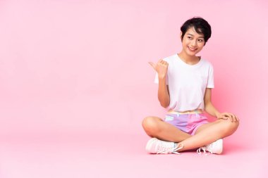 Kısa saçlı genç Vietnamlı kadın, izole edilmiş pembe arka planda yerde oturuyor ve bir ürünü sunmak için kenarı işaret ediyor.