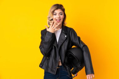 Sarı arka planda motosiklet kaskı tutan genç bir kadın. Sürpriz bir yüz ifadesi var.