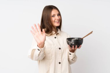 Beyaz arka planda izole edilmiş genç beyaz kız elinde çubuklarla elinde erişte kasesi tutarken mutlu bir ifadeyle selam veriyor.