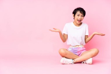 Kısa saçlı, genç Vietnamlı kadın, yüzü şoke olmuş bir halde pembe arka planda oturuyor.