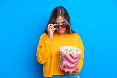 Mavi arka planda soyutlanmış beyaz bir kadın 3D gözlüklü ve elinde koca bir kova patlamış mısırla şaşırdı.