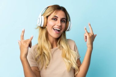 Genç sarışın Uruguaylı, mavi arka planda izole edilmiş rock hareketleri yaparak müzik dinliyor.