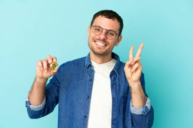 Brezilya 'lı bir adam mavi arka planda Bitcoin tutuyor gülümsüyor ve zafer işareti gösteriyor