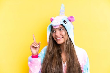 Sarı arka planda izole edilmiş tek boynuzlu at pijaması giyen beyaz bir kadın harika bir fikre işaret ediyor.