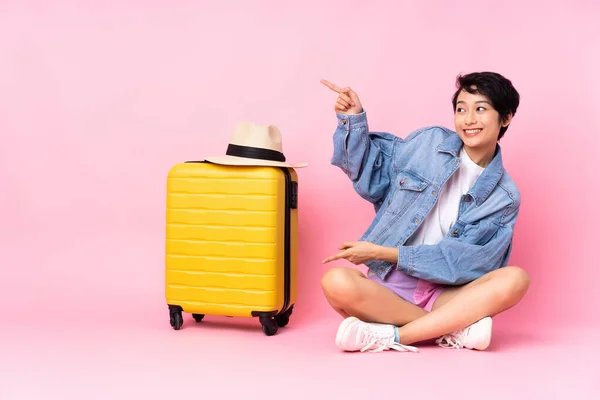 若いです旅行ベトナム人女性とともにスーツケース上の床の上に座って隔離されたピンクの背景指差し指に側と製品を提示 — ストック写真