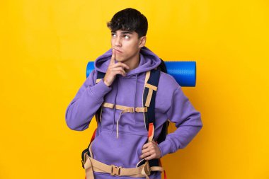 Sarı arka plan üzerinde büyük bir sırt çantası olan genç dağcı yukarı bakarken kuşkuları var.