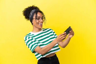 Sarı arka planda izole edilmiş genç Afrikalı Amerikalı kadın cep telefonuyla oynuyor.