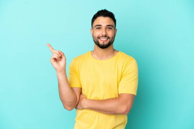 Genç Arap adam mavi arka planda izole edilmiş mutlu ve yukarıya bakıyor.