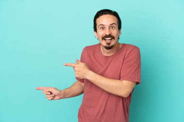 Mavi arka planda izole edilmiş genç beyaz adam şaşırmış ve işaret ediyor.