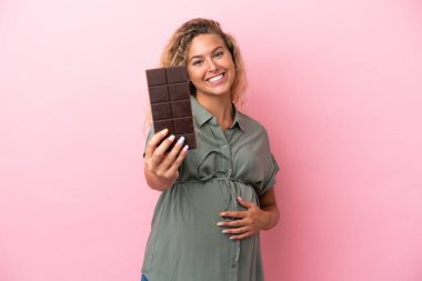 Kıvırcık saçlı kız pembe arka planda izole edilmiş hamile ve elinde çikolata var.