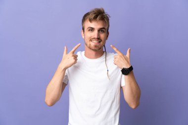 Mor arka planda izole edilmiş genç, yakışıklı sarışın bir adam baş parmak işareti yapıyor.