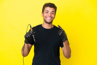 Dövme sanatçısı Brezilyalı adam sarı arka planda izole edilmiş bir şekilde baş parmak hareketi yapıyor.