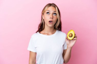 Avokado tutan genç Rus kadın pembe arka planda izole edilmiş yukarı bakıyor ve şaşırmış bir ifadeyle