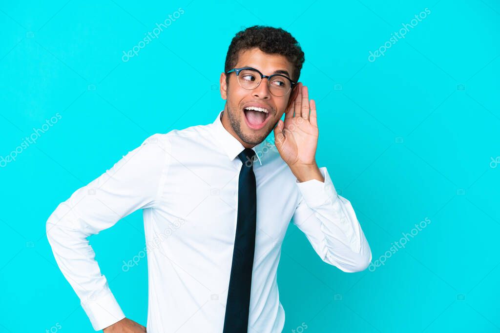 Young business Brazilian man isolated on blue background listening to something by putting hand on the ear