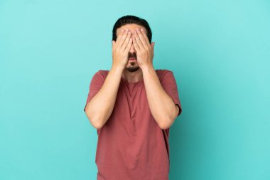 Genç beyaz adam mavi arka planda izole edildi. Gözlerini elleriyle kapadı.