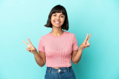 Mavi arka planda izole edilmiş genç melez kadın iki eliyle zafer işareti gösteriyor.