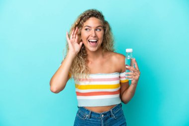 Mavi arka planda izole edilmiş bir şişe suyu olan genç sarışın kadın elini kulağına koyarak bir şey dinliyor.