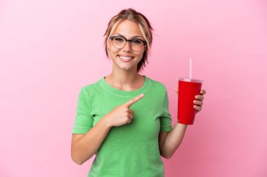 Pembe arka planda izole edilmiş bir içecek taşıyan genç Rus kadın bir ürünü sunmak için kenarı işaret ediyor.
