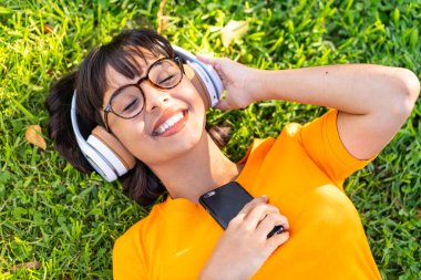 Genç esmer kadın açık havada cep telefonuyla müzik dinliyor.