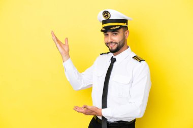 Arap uçak pilotu, sarı arka planda izole edilmiş ve davet edilmek için ellerini yana uzatıyor.