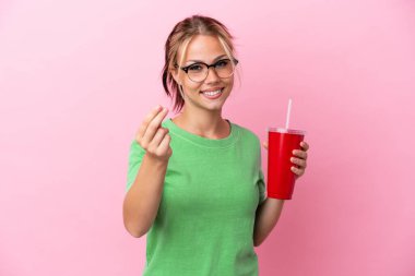 Genç Rus kadın pembe arka planda izole edilmiş bir içecek tutuyor ve para hareketi yapıyor.
