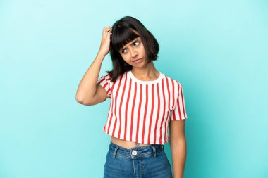 Genç melez kadın mavi arka planda izole edilmiş. Kafası kaşınırken şüpheleri var.