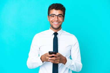Genç Brezilyalı iş adamı mavi arka planda izole edilmiş cep telefonuyla bir mesaj gönderiyor.
