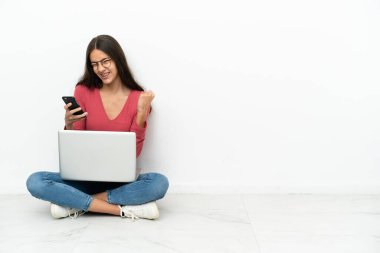 Genç Fransız kız dizüstü bilgisayarıyla yerde oturuyor ve telefonu zafer pozisyonunda.