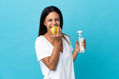 Beyaz kız mavi arka planda bir şişe suyla izole edilmiş ve elma yiyor.