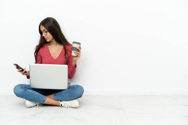 Genç bir Fransız kız laptopuyla yerde oturuyor elinde kahve ve cep telefonu var.