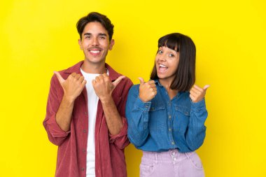 Sarı arka planda izole edilmiş genç melez bir çift iki eliyle baş parmak işareti yapıyor ve gülümsüyor.