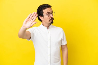 Sarı arka planda izole edilmiş genç beyaz adam dur hareketi yapıyor ve hayal kırıklığına uğruyor.