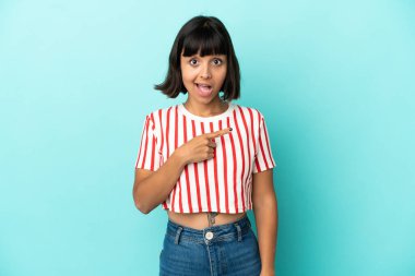 Mavi arka planda izole edilmiş genç melez kadın şaşırmış ve işaret ediyor.