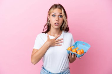 Pembe arka planda yumurtaları tutan genç Rus kadın şaşırdı ve şaşırdı.