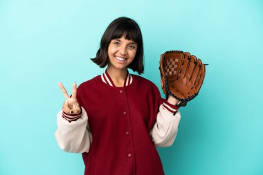 Beyzbol eldivenli genç melez kadın mavi arka planda izole edilmiş gülümsüyor ve zafer işareti gösteriyor.