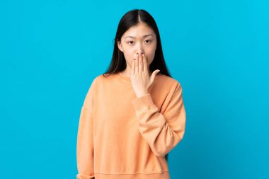 Genç Çinli kız, izole edilmiş mavi arka planda ağzını eliyle kapatıyor.