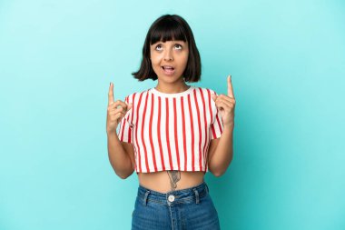 Mavi arka planda izole edilmiş genç melez kadın şaşırmış ve işaret ediyor.