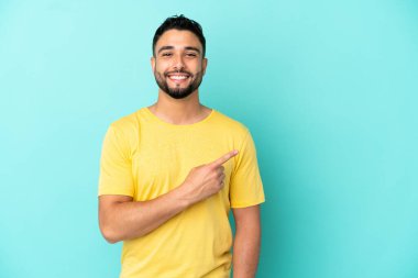 Mavi arka planda izole edilmiş genç Arap bir ürünü sunmak için kenarı işaret ediyor.