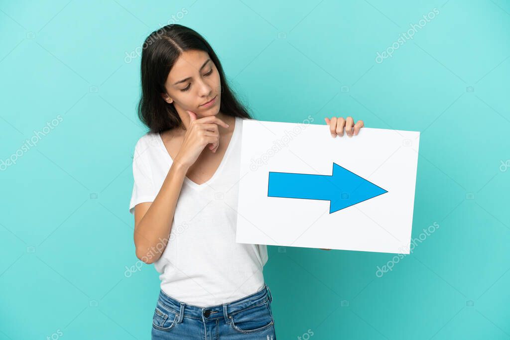 Young French woman isolated on blue background holding a placard with arrow symbol and thinking