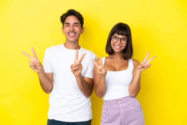 Sarı arka planda izole edilmiş genç melez çift gülümsüyor ve zafer işareti gösteriyorlar.